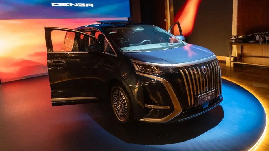Vista frontal do veículo elétrico Denza Z9 GT em exposição, com portas de motorista e passageiro abertas, em um ambiente de estúdio com iluminação azul e painel ao fundo.